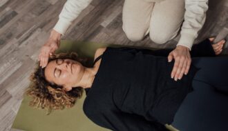 A practitioner kneels beside a person lying on a mat, hands held just above her forehead and chest during a Reiki session