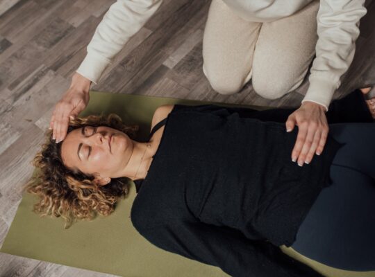 A practitioner kneels beside a person lying on a mat, hands held just above her forehead and chest during a Reiki session