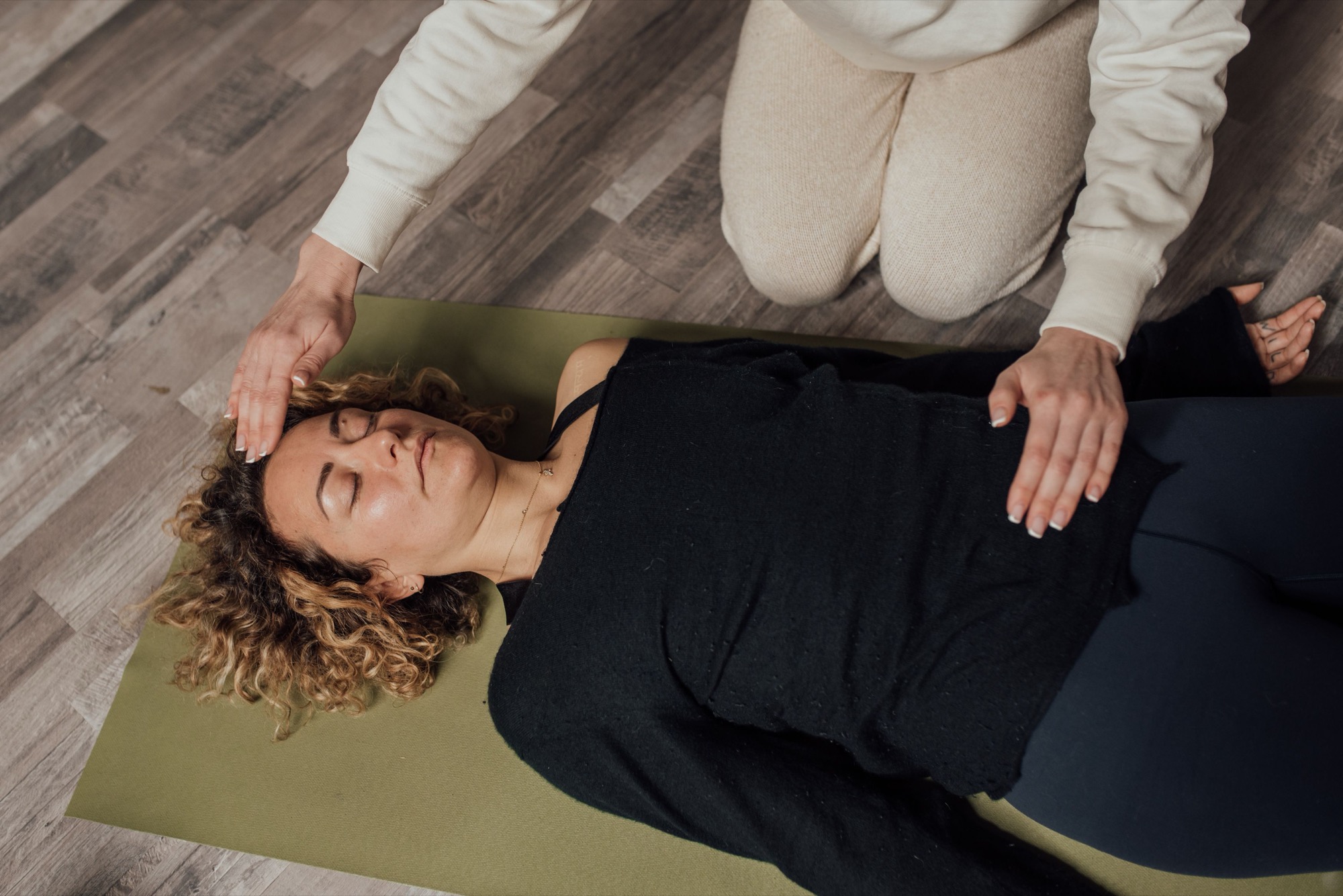 A practitioner kneels beside a person lying on a mat, hands held just above her forehead and chest during a Reiki session